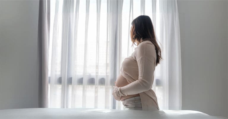Caucasian pregnant woman staring out a window with white curtains
