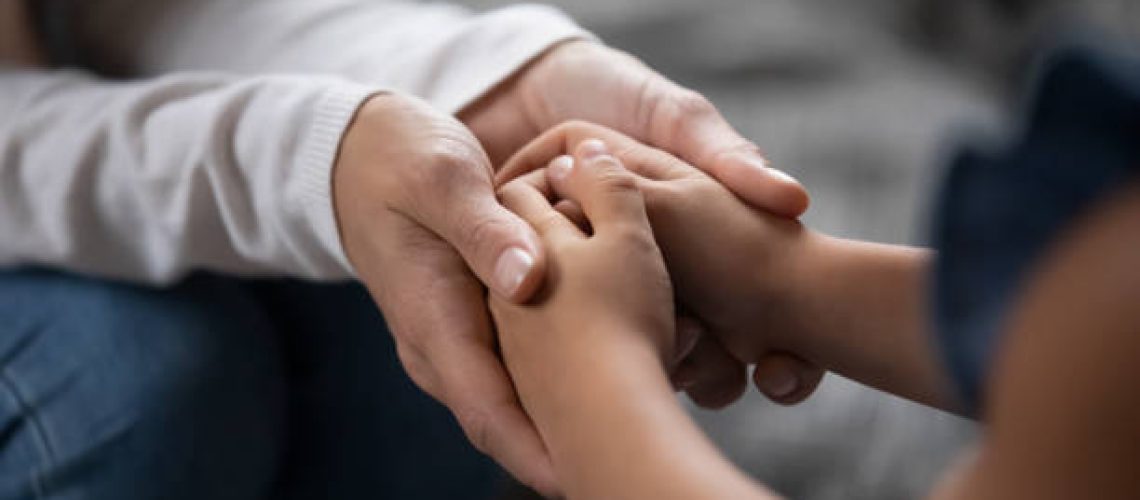 Close up compassionate young foster parent holding hands of little kid girl, giving psychological help, supporting at home. Sincere different generations family sharing secrets or making peace.